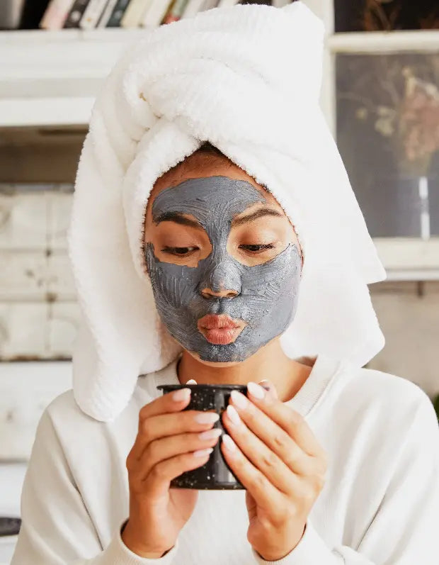 Person applying a facial mask with a towel on their head, holding a black container.
