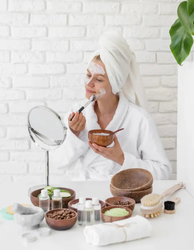 Woman applying facial mask in front of a mirror with various skincare products on a white brick wall background.