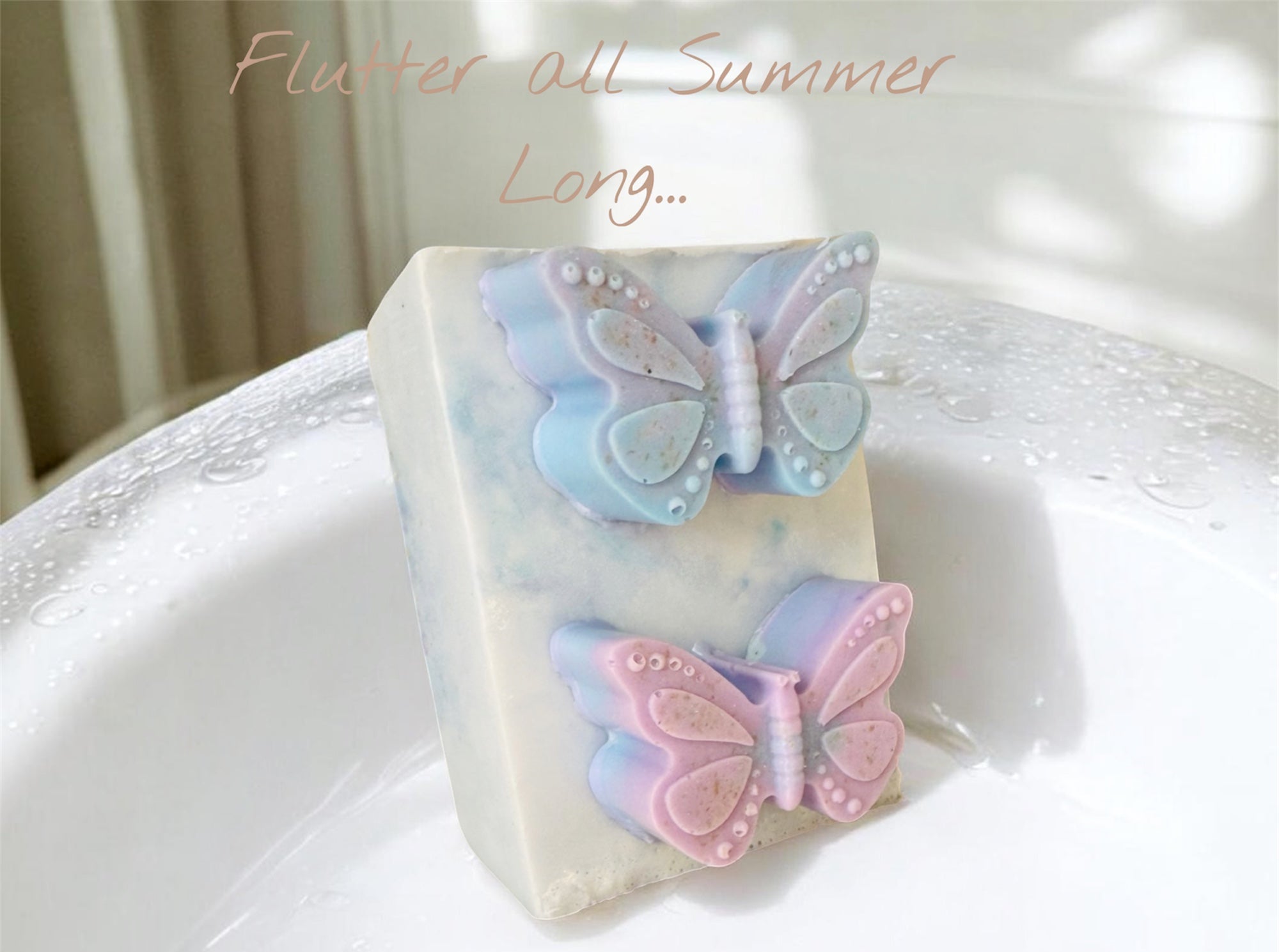 A rectangular bar of soap with two butterfly decorations resting in a white sink. The top butterfly is light blue and lavender, and the bottom butterfly is pink and lavender.