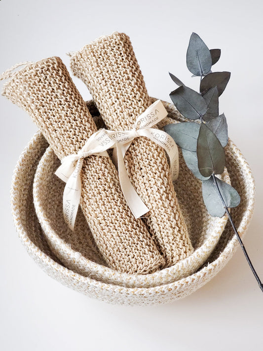 Three rolled hemp washcloths tied with ribbon in a woven basket beside a eucalyptus stem